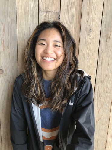 A young woman with wavy hair, wearing a black jacket and a blue t-shirt, smiling in front of a wooden background.